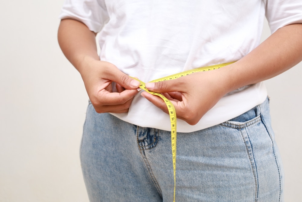 Close-up of a woman in a white shirt measuring her waist with a yellow tape measure, symbolizing weight loss, body slimming, calorie tracking, and a successful healthy lifestyle or diet plan.
