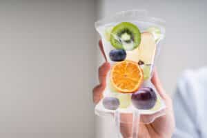 Hand holds an IV therapy bag aloft, with images of an orange, kiwi, and grapes appearing to represent the nutrients it offers