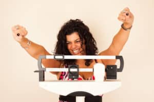 Woman with brown curly hair stands on a scale and cheers after medical weight loss with habit tracking